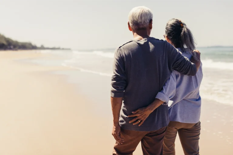 Pareja mayor abrazada de espaldas contemplando el mar en una playa soleada.
