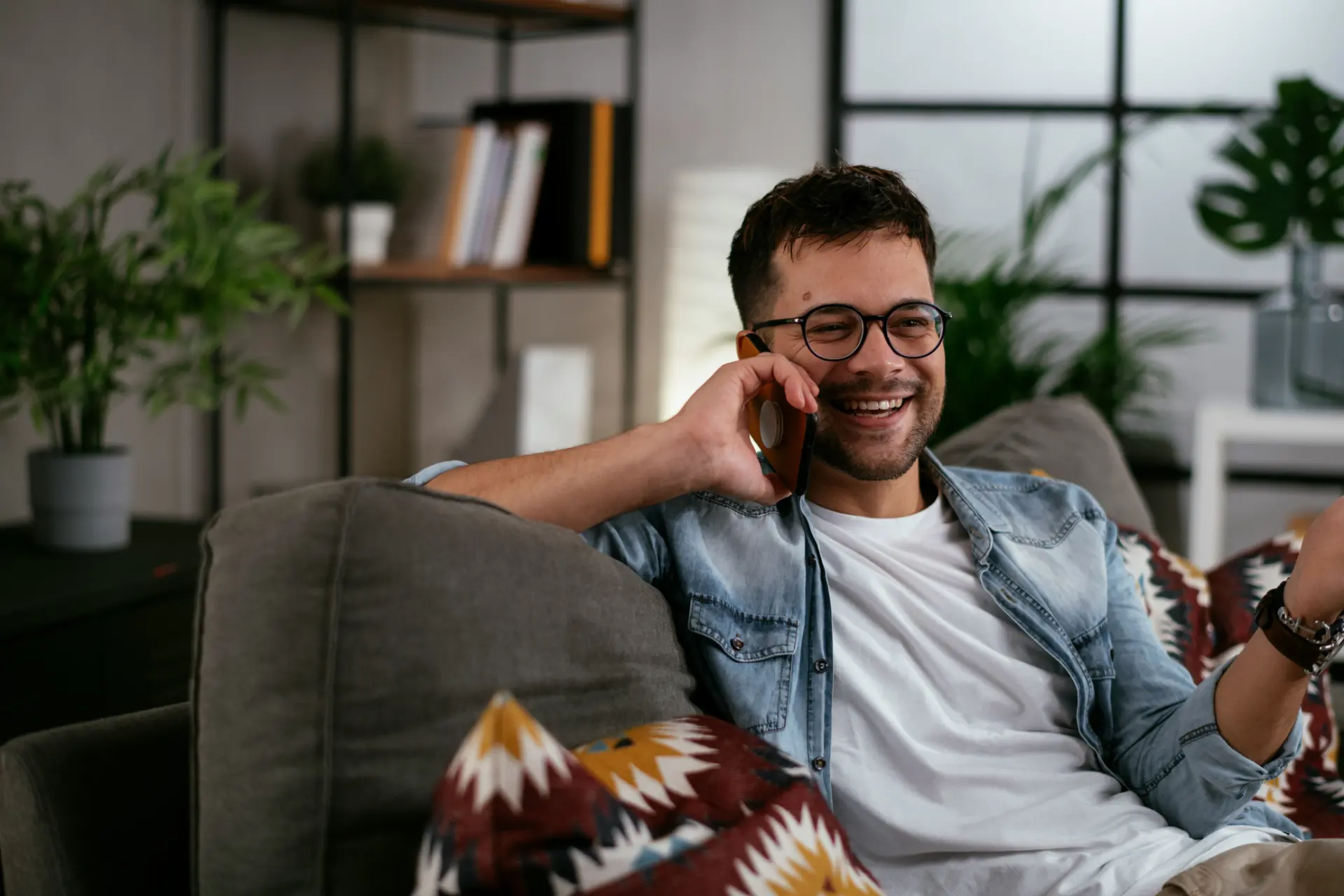 Hombre joven con gafas sonriendo mientras habla por teléfono relajado en el sofá de su hogar.