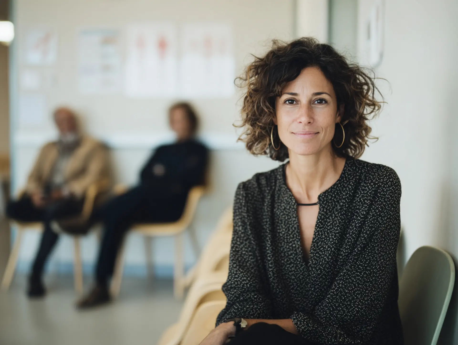 Mujer española serena sentada en una sala de espera médica con otros pacientes al fondo.