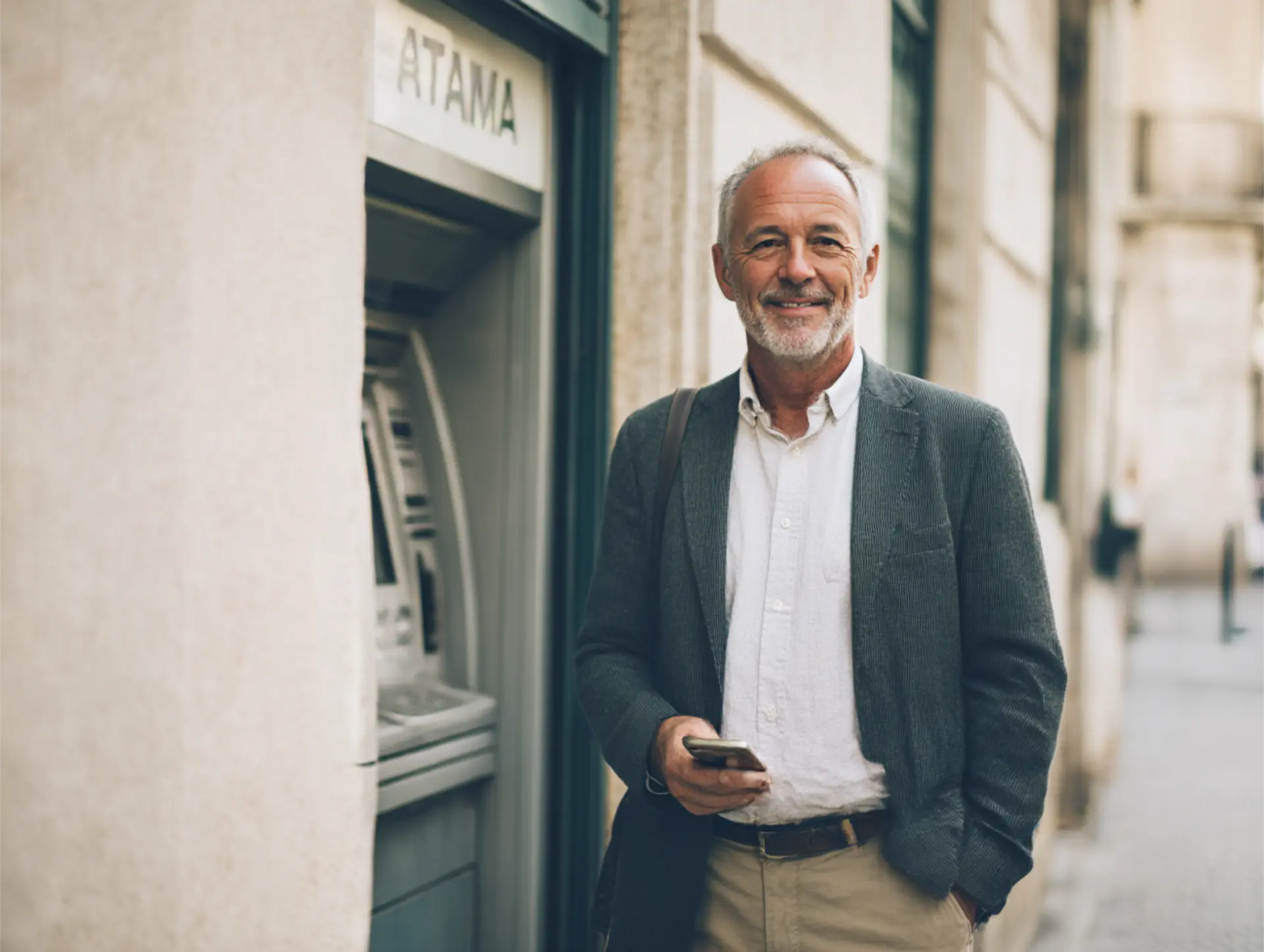 Hombre mayor sonriendo junto a un cajero automático en la calle con el móvil en la mano.