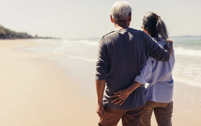 Pareja mayor abrazada de espaldas contemplando el mar en una playa soleada.