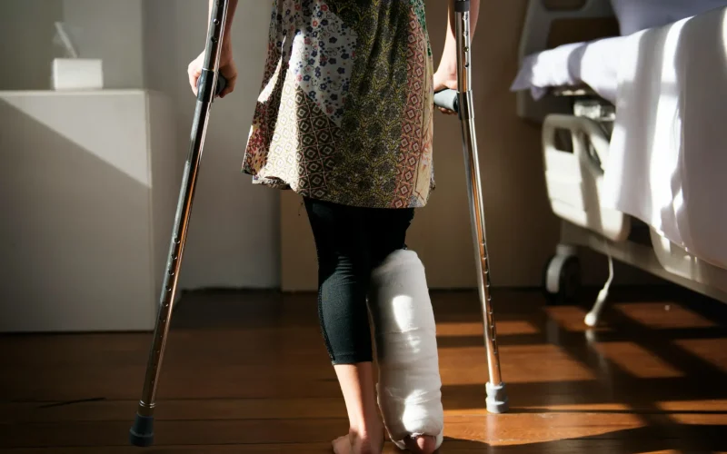 Mujer con escayola en el pie caminando con muletas en una habitación de hospital iluminada.
