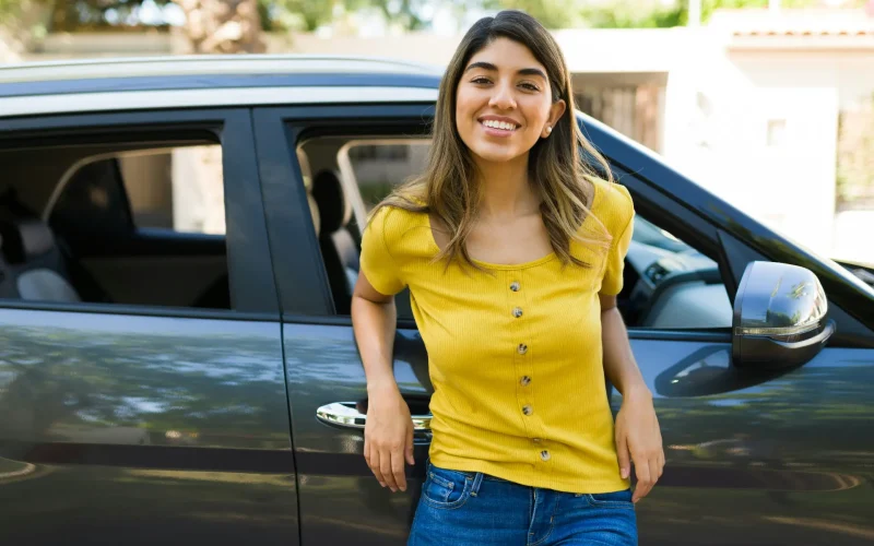 Mujer joven con camiseta amarilla sonriendo apoyada en la puerta de su coche.