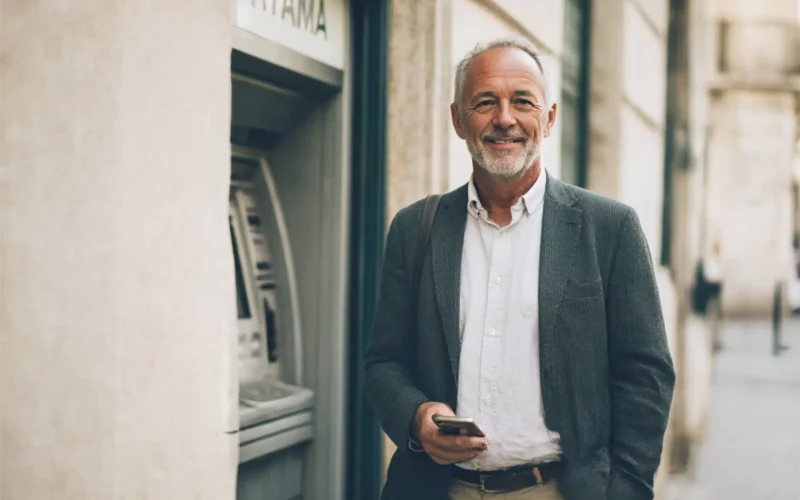 Hombre mayor sonriendo junto a un cajero automático en la calle con el móvil en la mano.